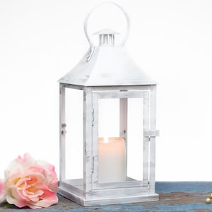 Puede incluir: Linterna de metal blanco con una vela en su interior. La linterna tiene un acabado blanco desgastado y está sentada sobre una superficie de madera con una rosa rosa.