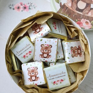 Può includere: Una scatola rotonda bianca piena di cioccolatini confezionati singolarmente. Ogni involucro di cioccolato presenta un orsetto dei cartoni animati con elementi floreali e il testo "Annaviola Prima Comunione". La scatola è foderata con carta marrone.