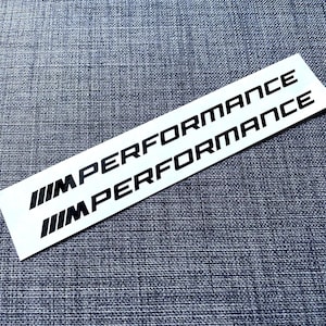Puede incluir: Dos pegatinas rectangulares blancas con el texto "M PERFORMANCE" en negro, apiladas una encima de la otra. Las pegatinas también presentan tres líneas negras en ángulo en el lado izquierdo. Sobre una superficie gris texturizada.