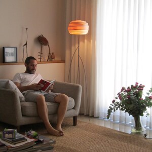 May include: A man sits in a gray armchair reading a book. He is wearing a white t-shirt and gray shorts. A large, round, layered lamp with a wooden shade sits on a tripod stand in the background. A bouquet of pink flowers sits in a clear glass vase on the floor.