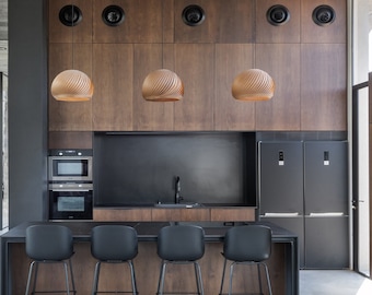 May include: A modern kitchen with a large island featuring a black countertop and four black bar stools. The kitchen has a wood-paneled wall with four pendant lights and a black range hood. The kitchen is open to a living area with large windows.
