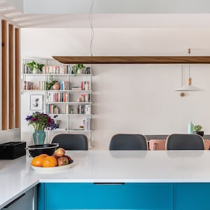 Può includere: Un piano di lavoro da cucina bianco con un mobiletto blu sottostante. Sul piano di lavoro c'è una ciotola di frutta. Lo sfondo mostra una sala da pranzo con una parete bianca, una mensola in legno con libri e una lampada a sospensione.