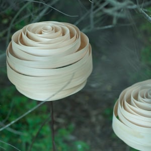 May include: Three wooden rose sculptures made of thin strips of wood, arranged in a spiral pattern, are mounted on metal stems. The sculptures are displayed against a background of green foliage.