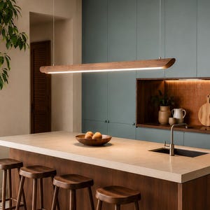 May include: A long, linear wooden pendant light fixture suspended by thin wires above a kitchen island. The fixture features a smooth, curved dark wood exterior with an integrated LED light strip running along the bottom edge, providing illumination.