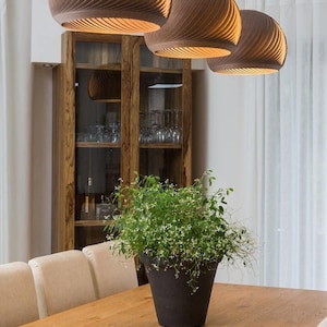 May include: A wooden dining table with a light wood finish. Three brown, woven pendant lights hang above the table. A potted plant sits in the center of the table.