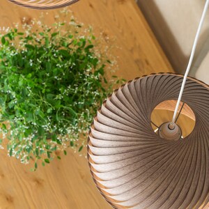 K&ouml;nnte beinhalten: Zwei Holz-Pendelleuchten mit einem einzigartigen Spiraldesign h&auml;ngen von der Decke &uuml;ber einem Holztisch. Die Leuchten bestehen aus d&uuml;nnen Holzstreifen, die &uuml;bereinander gelegt und gebogen sind, um eine Spiralform zu erzeugen. Die Leuchten sind hellbraun und haben eine nat&uuml;rliche Holzoptik.