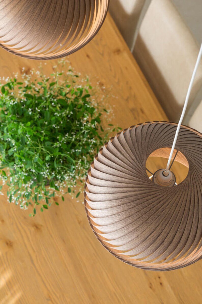 K&ouml;nnte beinhalten: Zwei Holz-Pendelleuchten mit einem einzigartigen Spiraldesign h&auml;ngen von der Decke &uuml;ber einem Holztisch. Die Leuchten bestehen aus d&uuml;nnen Holzstreifen, die &uuml;bereinander gelegt und gebogen sind, um eine Spiralform zu erzeugen. Die Leuchten sind hellbraun und haben eine nat&uuml;rliche Holzoptik.