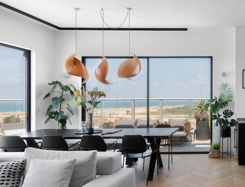 K&ouml;nnte beinhalten: Ein modernes Esszimmer mit einem Holztisch und schwarzen St&uuml;hlen. Der Raum hat gro&szlig;e Fenster mit Blick auf das Meer und einen Balkon. Drei h&ouml;lzerne Pendelleuchten h&auml;ngen &uuml;ber dem Tisch.