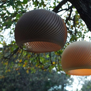 May include: Two round, hanging, woven light fixtures with a wood grain pattern. The top fixture is brown and the bottom fixture is beige. The fixtures are hanging from a tree branch.