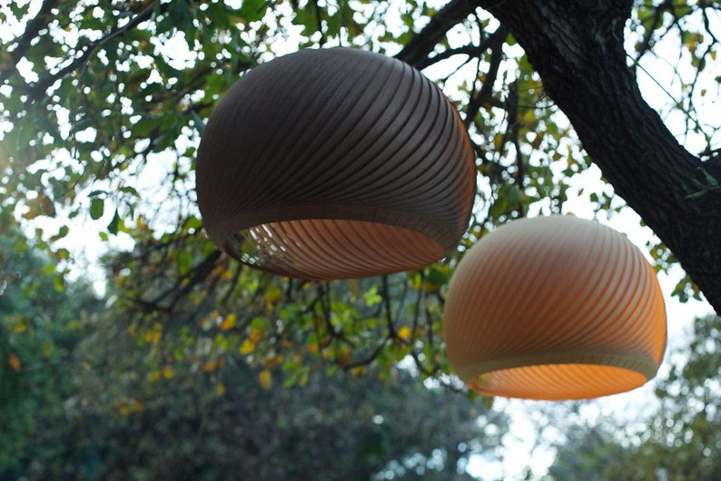 May include: Two round, hanging, woven light fixtures with a wood grain pattern. The top fixture is brown and the bottom fixture is beige. The fixtures are hanging from a tree branch.