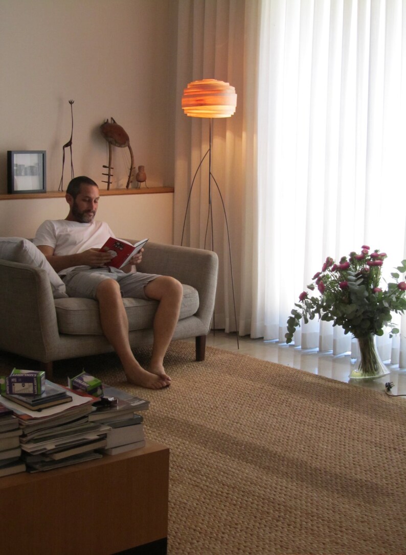 May include: A man sits in a gray armchair reading a book. He is wearing a white t-shirt and gray shorts. A large, round, layered lamp with a wooden shade sits on a tripod stand in the background. A bouquet of pink flowers sits in a clear glass vase on the floor.