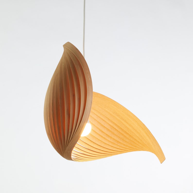 May include: A modern pendant light fixture with a leaf-like design. The light is made of light-colored wood and features a bulb in the center. The fixture is suspended from a white cord against a white background.