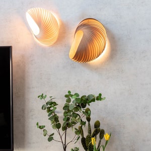 May include: Two wooden wall lights with a geometric design, illuminated with warm white light, mounted on a light grey wall. A metal tray with potted plants, including a cactus, a succulent, and a leafy plant, sits on a light grey surface.