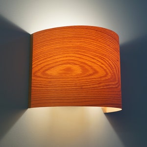 May include: A curved wooden wall sconce with a warm light emanating from it. The sconce has a wood grain pattern and is mounted on a wall.