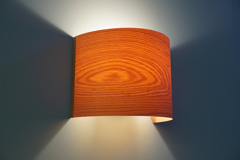 May include: A curved wooden wall sconce with a warm light emanating from it. The sconce has a wood grain pattern and is mounted on a wall.