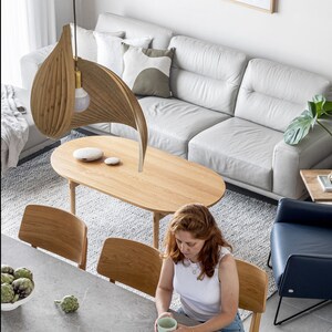May include: An overhead shot of a modern living space with a wooden oval coffee table, a light gray sofa, and a unique wooden leaf-shaped pendant light. A person is seated at a gray countertop table with a cup. The room has a minimalist design.