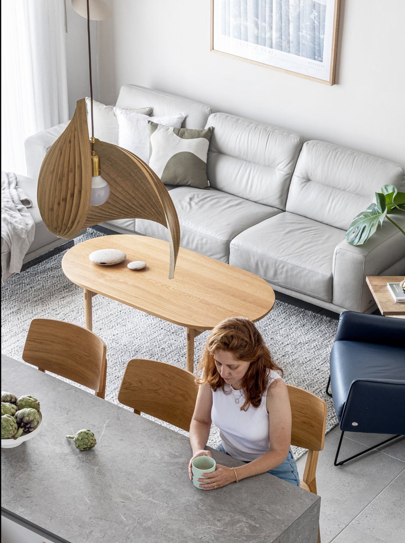 May include: An overhead shot of a modern living space with a wooden oval coffee table, a light gray sofa, and a unique wooden leaf-shaped pendant light. A person is seated at a gray countertop table with a cup. The room has a minimalist design.