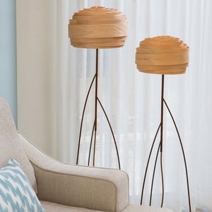 May include: Two wooden floor lamps with a layered, rounded shade. The lamps have a dark brown metal base with a tripod design. The lamps are in front of a white sheer curtain.