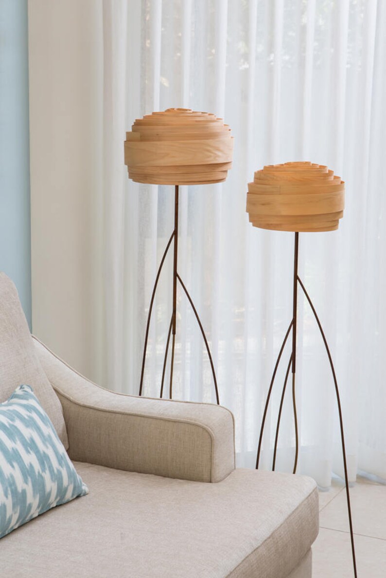 May include: Two wooden floor lamps with a layered, rounded shade. The lamps have a dark brown metal base with a tripod design. The lamps are in front of a white sheer curtain.