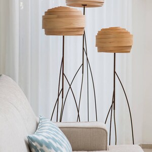 May include: Three wooden floor lamps with layered shades in a living room setting. The lamps have a modern design with thin black metal bases.
