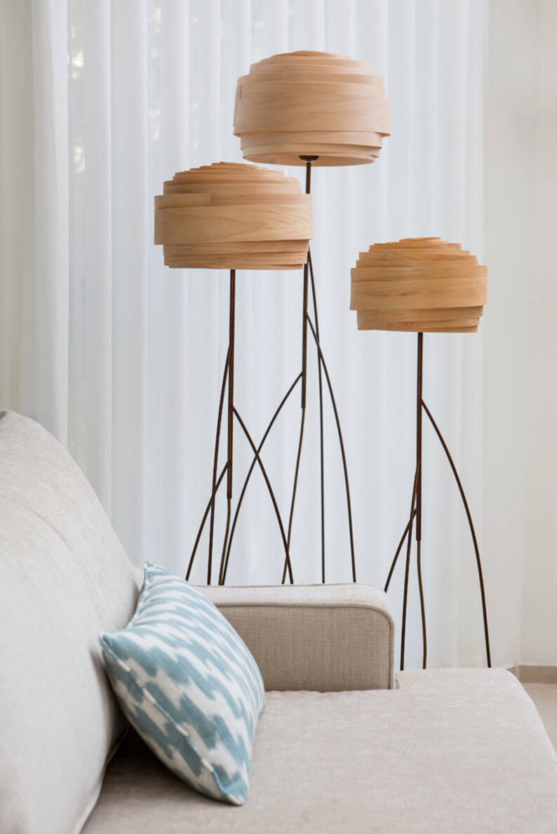 May include: Three wooden floor lamps with layered shades in a living room setting. The lamps have a modern design with thin black metal bases.