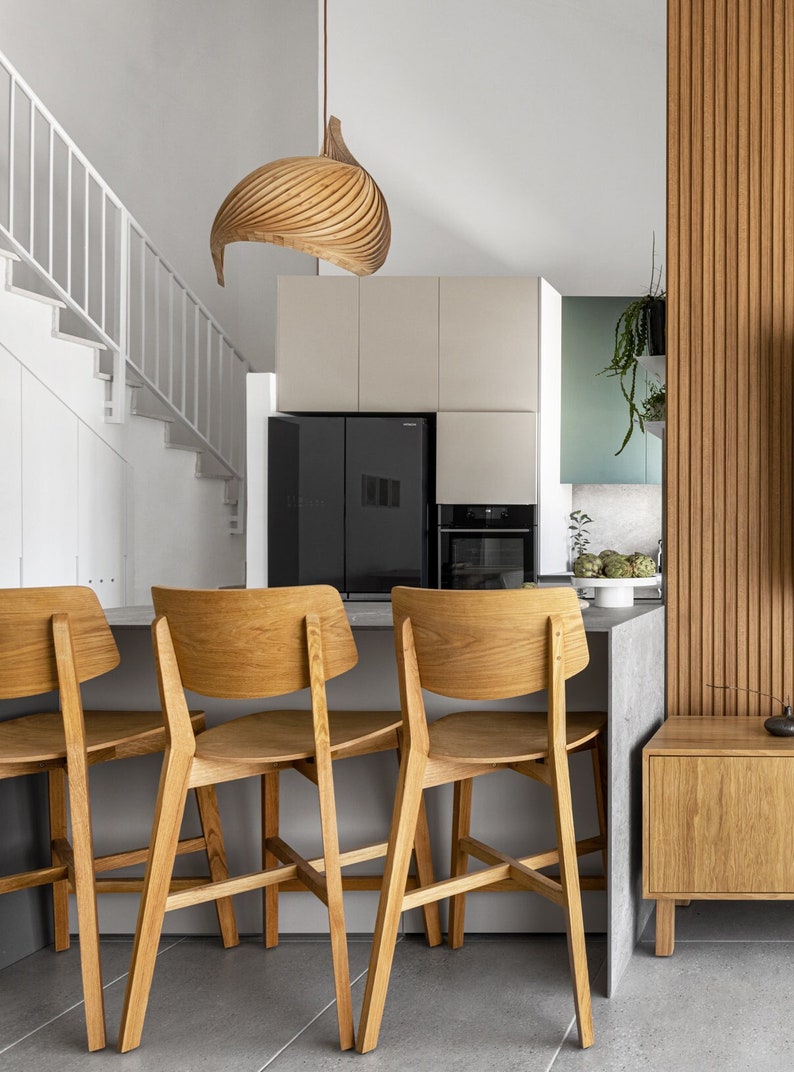 May include: A modern kitchen interior featuring three wooden bar stools with light-colored wood. A black refrigerator and oven are set against a neutral-toned cabinet. A wooden pendant light hangs above the bar. The kitchen has a minimalist design.