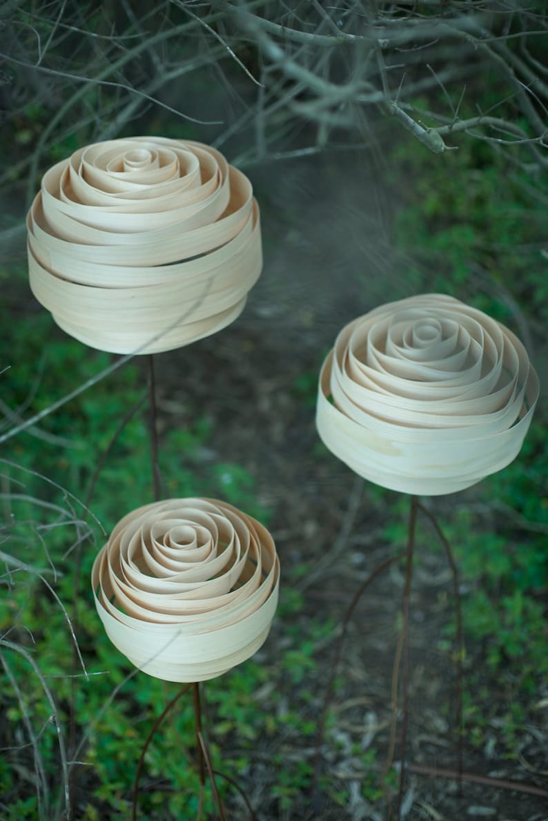 May include: Three wooden rose sculptures made of thin strips of wood, arranged in a spiral pattern, are mounted on metal stems. The sculptures are displayed against a background of green foliage.