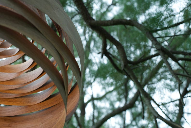 May include: A close-up of a wooden lampshade with a spiral design. The lampshade is made of thin strips of wood that are woven together. The lampshade is hanging from the ceiling and is partially obscured by a tree branch.