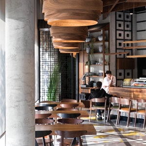 May include: A modern restaurant interior with wooden tables and chairs. The restaurant has a minimalist design with a concrete wall and wooden ceiling beams. The tables are arranged in rows and are illuminated by large, wooden pendant lights.