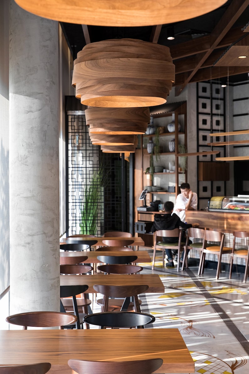 May include: A modern restaurant interior with wooden tables and chairs. The restaurant has a minimalist design with a concrete wall and wooden ceiling beams. The tables are arranged in rows and are illuminated by large, wooden pendant lights.