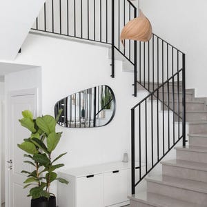 May include: Interior shot of a modern staircase with gray steps and black metal railings. A large potted plant sits near a white cabinet and a unique, irregularly shaped mirror. Two wooden pendant lights hang above.