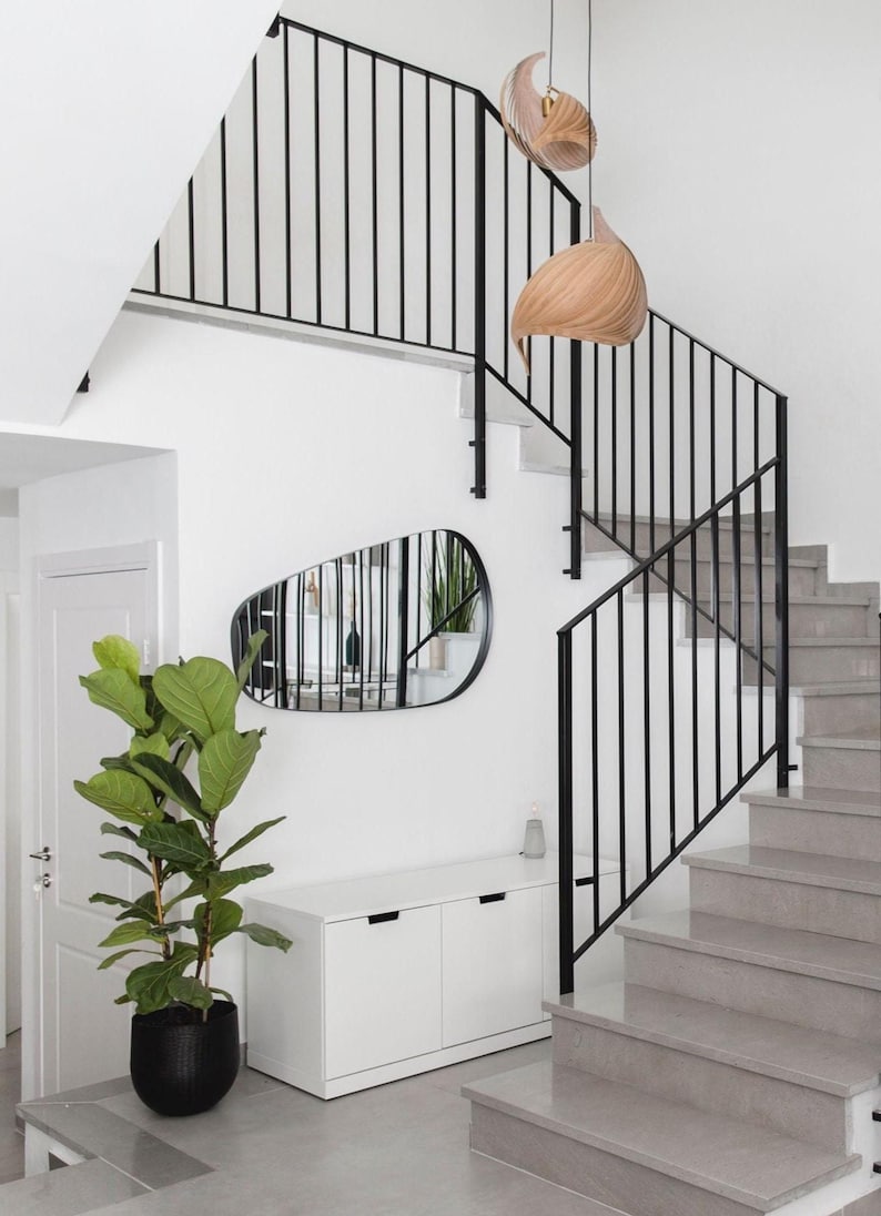 May include: Interior shot of a modern staircase with gray steps and black metal railings. A large potted plant sits near a white cabinet and a unique, irregularly shaped mirror. Two wooden pendant lights hang above.