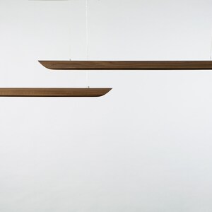 Op de afbeelding: Twee houten hanglampen die aan het plafond hangen. De lampen zijn lang en smal, met een licht gebogen vorm. Ze zijn gemaakt van donker hout.