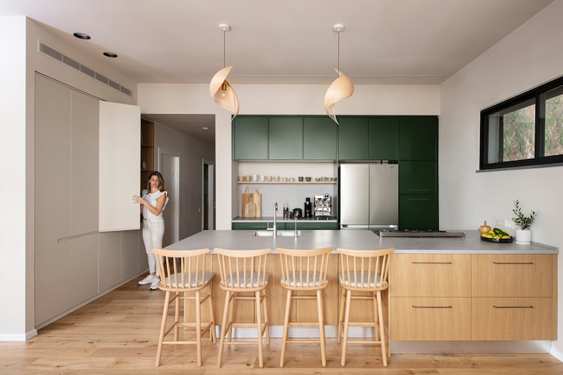 K&ouml;nnte beinhalten: Eine moderne K&uuml;che mit gr&uuml;nen Schr&auml;nken, einem K&uuml;hlschrank aus Edelstahl und einer gro&szlig;en Insel mit vier Holzbarhockern. Die Insel hat eine hellgraue Arbeitsplatte und eine Holzbasis. Die K&uuml;che hat einen Holzboden und ein gro&szlig;es Fenster mit Blick nach drau&szlig;en.