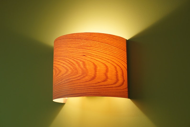 May include: A wooden wall sconce with a cylindrical shade. The shade has a wood grain pattern and is lit from within, casting a warm glow.