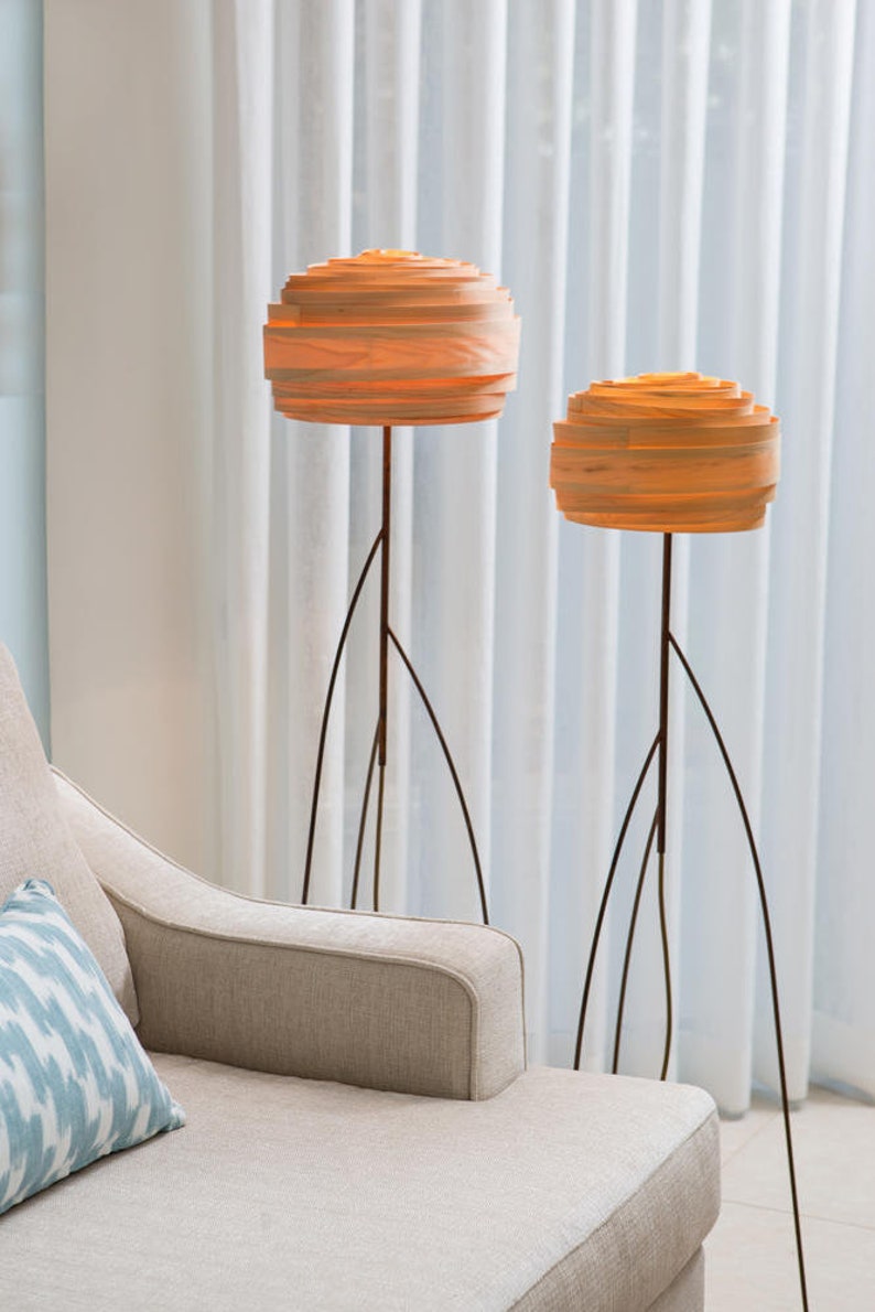 May include: Two modern floor lamps with wooden shades and thin black metal stands. The lamps are lit and casting a warm glow. The lamps are in front of a white sheer curtain.