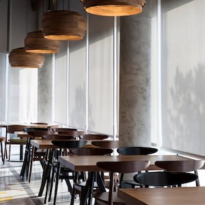 May include: A modern dining area with wooden tables and chairs. The tables are rectangular and the chairs are black with a curved back. The room has large windows with white blinds and wooden pendant lights.
