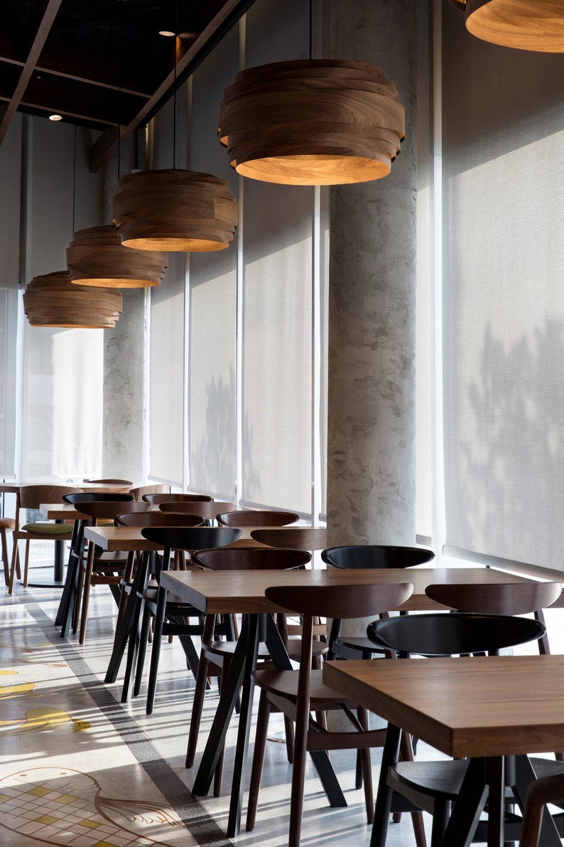 May include: A modern dining area with wooden tables and chairs. The tables are rectangular and the chairs are black with a curved back. The room has large windows with white blinds and wooden pendant lights.