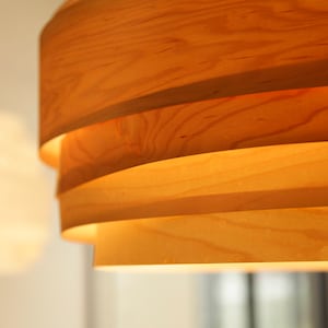 Könnte beinhalten: Nahaufnahme einer Holz-Pendelleuchte mit mehreren Holzschichten. Die Leuchte ist aus Naturholz gefertigt und strahlt ein warmes, einladendes Licht aus.