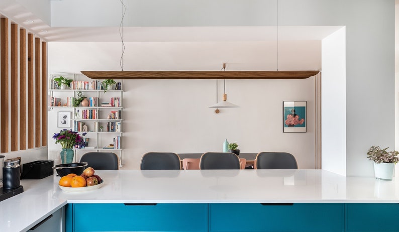 Op de afbeelding: Een wit keukenblad met daaronder een blauwe kast. Op het blad staat een kom met fruit. Op de achtergrond is een eetkamer te zien met een witte muur, een houten boekenplank en een hanglamp.