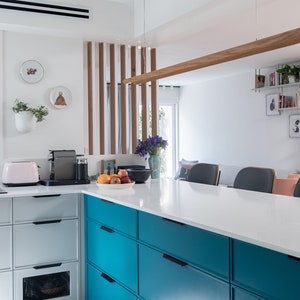 Op de afbeelding: Een moderne keuken met witte kasten en een turquoise eiland met laden. Het eiland heeft een wit aanrechtblad en een houten lamp die erboven hangt. Er is een boekenkast op de achtergrond met boeken en planten.