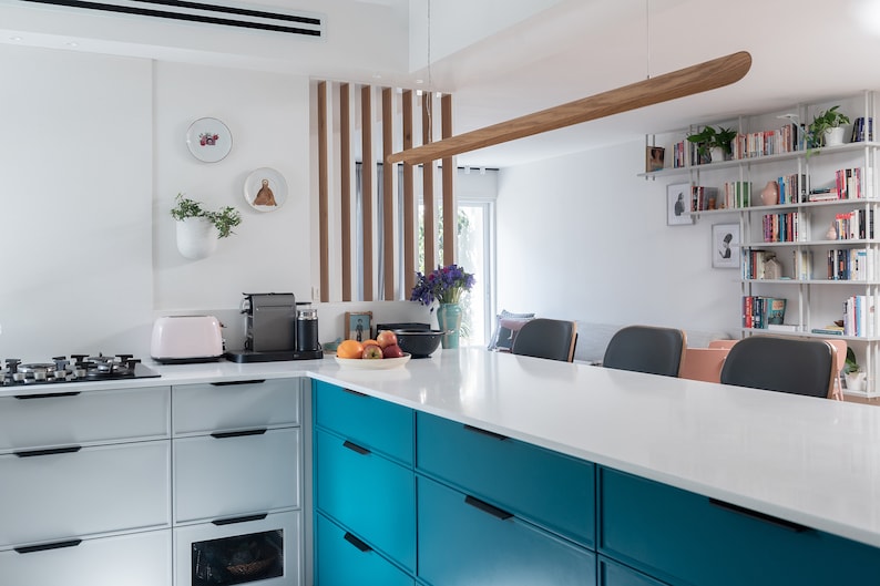 Op de afbeelding: Een moderne keuken met witte kasten en een turquoise eiland met laden. Het eiland heeft een wit aanrechtblad en een houten lamp die erboven hangt. Er is een boekenkast op de achtergrond met boeken en planten.