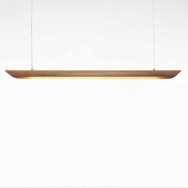 Op de afbeelding: Een lange, smalle, houten hanglamp met een warme, gouden gloed. De lamp wordt aan het plafond gehangen met een dun koord.