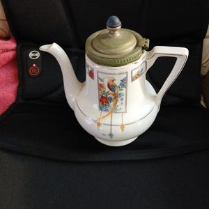 May include: White porcelain teapot with a decorative bird and floral design. The teapot has a metal lid and a blue knob. The handle and spout are white. The teapot is a vintage item.