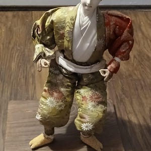 May include: A Japanese-style doll with a pale face and black hair tied in a topknot. The doll wears a white undergarment, a green patterned kimono with floral motifs, and a contrasting red sleeve on the left arm, standing on a square wooden base.