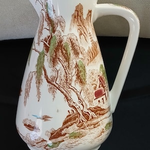 May include: White ceramic pitcher with a brown and green landscape design. The pitcher features a curved handle and a spout. The landscape includes trees, mountains, and a small house. The design is reminiscent of vintage tableware.