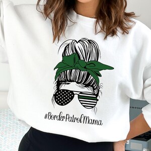 May include: White sweatshirt with a graphic of a woman's updo hairstyle, green headband, and sunglasses with an American flag design. The text "#Border Patrol Mama" is below the graphic.