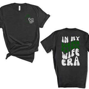 May include: A dark grey t-shirt with a small heart and flower design on the left chest. The back of the shirt has the text "IN MY WIFE ERA" in a green and white retro font.