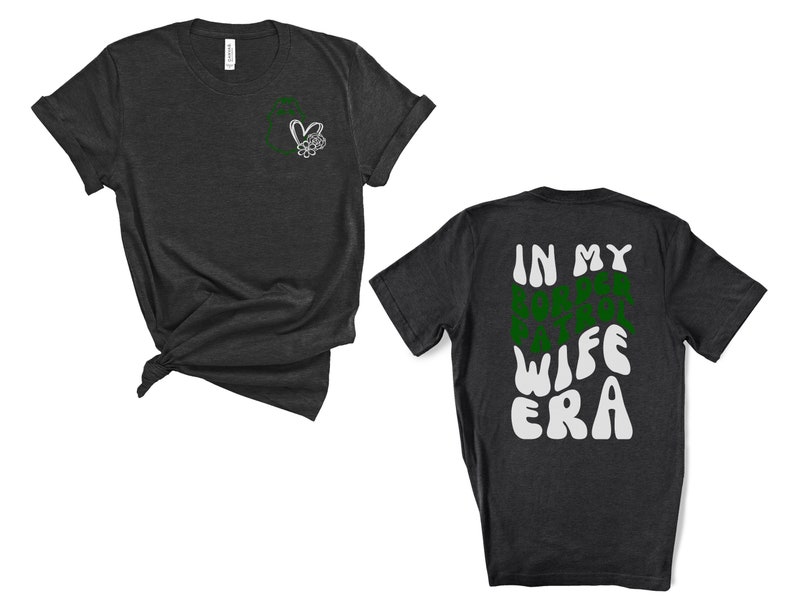 May include: A dark grey t-shirt with a small heart and flower design on the left chest. The back of the shirt has the text "IN MY WIFE ERA" in a green and white retro font.