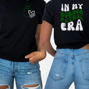 May include: A black t-shirt featuring a small green badge outline and white heart-flower graphic on the front left chest. The back displays the text IN MY BORDER PATROL ERA in large, wavy white and green lettering on a black background.
